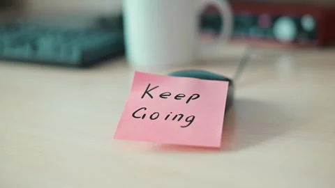 A pink sticky note with the message Keep Going is placed on a table. A white mug Stock Footage 339094045