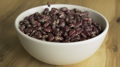 Pinto beans falling into white bowl on wooden table 4k 10bit 120fps Видео 333352722