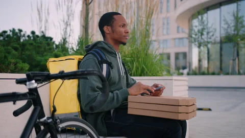 Pizza deliveryman is sitting on a bench with a mobile phone while waiting Stock Footage 222889716