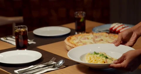 Placing pasta on table set with pizza and drinks. Cozy dining scene in Видео 311998637