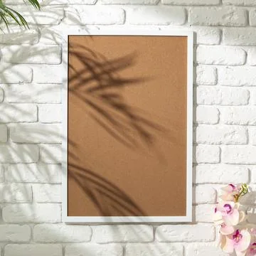 Plain cork board is mounted on a white brick wall with pink flowers nearby in a Stock Photos