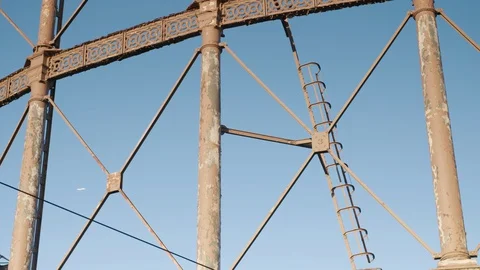 A plane passes behind an ornate rusting Victorian gas holder with ladder at 스톡 동영상 128920929