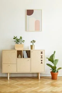 Plant next to wooden cupboard against white wall with poster in living room i Stock Photos
