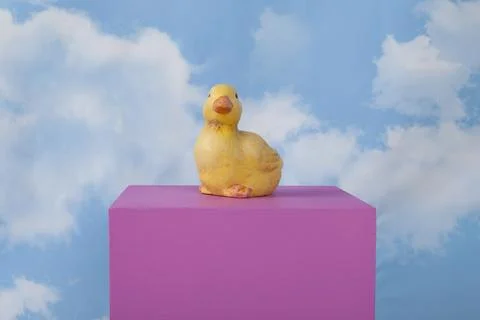 A plaster duck on a pink cube in front of a blue sky with clouds. Stock Photos