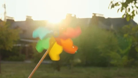 Plastic rainbow windmill rotates on the background of blurry houses and sunset Stock Footage 119889982