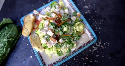 Plate of delicious salad surrounded by vegetables and salt on black surface Stock Footage 168640752