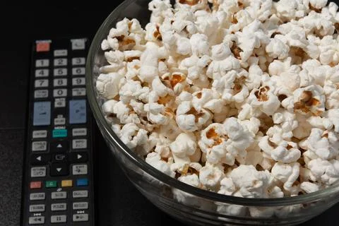 A plate of popcorn and a TV remote on a black background. Stock Photos