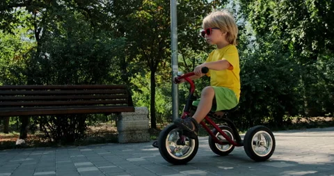 Playful child in bright clothes rides tricycle past bushes Stock Footage