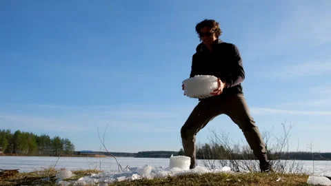 Playful man breaks a block of ice near frozen river on nature in sunny day. Stock Footage 311860082