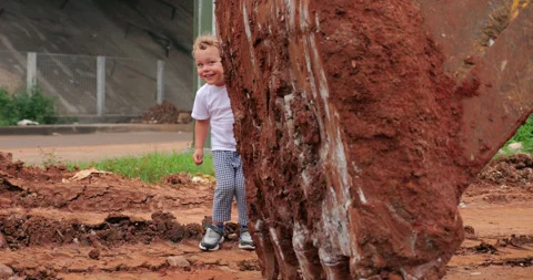 Playful tiny boy hides and peeks with smile from behind bucket of excavator Video stock 278475391