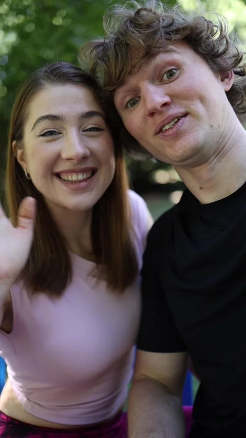 Playful young couple wave at camera while filming a cheerful selfie in park Vídeos de archivo 341476755