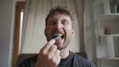 Playful young man with a beard sitting indoors eating colorful gummy dinosaur Stock Footage 335471268