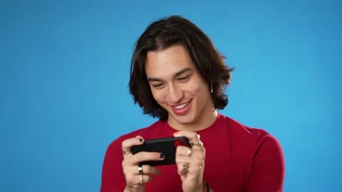 Playing game on phone smiling Hispanic Latino gender fluid young man 20s in red Stock Footage 220775617