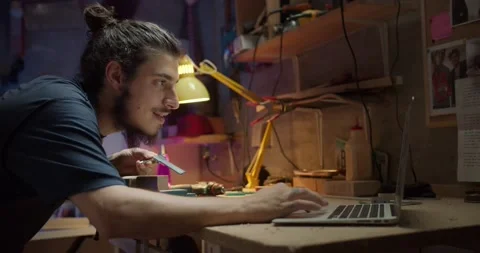 Pleasant artisan male using computer online while making guitar arm in evening Stock Footage 137242814