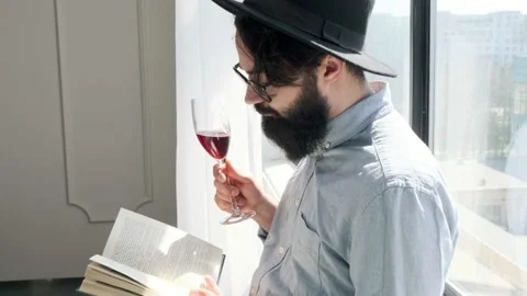 Pleased bearded man drinking red wine while reading a book at home. Stock Footage 152837108