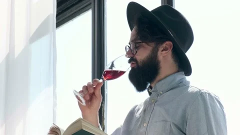 Pleased bearded man drinking red wine while reading a book at home. Stock Footage 153482390