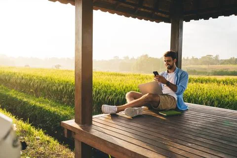 Pleased man browsing smartphone while using laptop for telework on terrace Foto stock