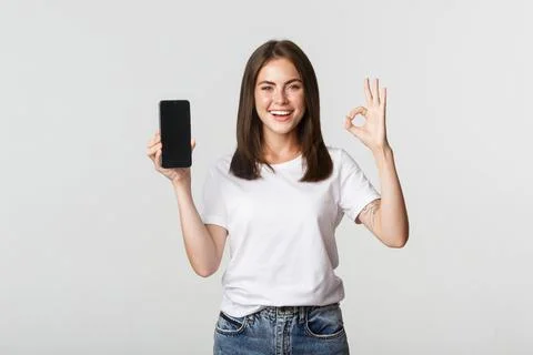 Pleased smiling brunette girl showing smartphone screen and showing okay gesture Stock Photos