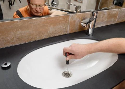 The plumber with a screwdriver fixes the sink drain in the bathroom. Stock Photos