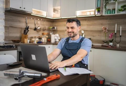 Plumber working on laptop in kitchen looking at instructions for repairing Stock Photos