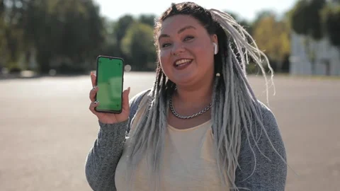 Plump cheerful woman with dreadlocks points at a phone with a green screen with Stock Footage 136544636