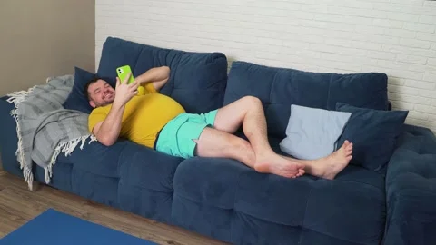 Plump man is doing physical exercise. Lying on the couch, communicating via Stock Footage 170608864