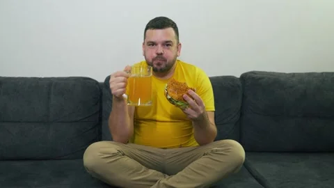 Plump young man is sitting on gray sofa and eating big fat burger and drinking Stock Footage 166933239