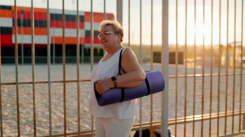 Plus size elder carrying yoga mat walking by fence at sunrise. fitness and 库存影片 340994230