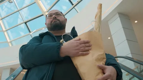 Plus size man on escalator with diet food in eco package in shopping mall Video stock 279647467