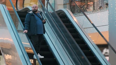 Plus size man standing on escalator with smartphone in shopping mall, showcasing Stock Footage 285537289