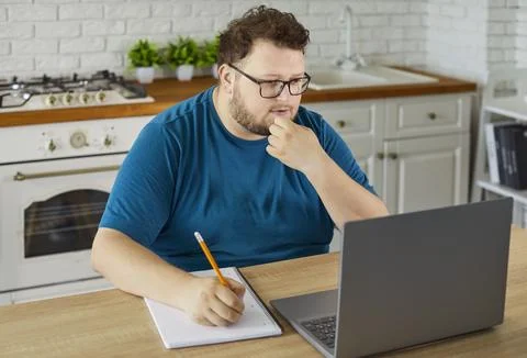 Plus size man studying online at home kitchen with laptop, notebook and pencil Stock Photos