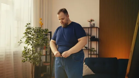 Plus-size man trying on jeans near mirror, happy with weight loss progress Stock Footage 308465584