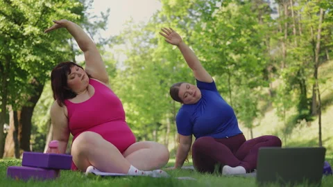 Plus-size sisters watching an online tutorial on laptop and exercising in park 動画素材 333633999
