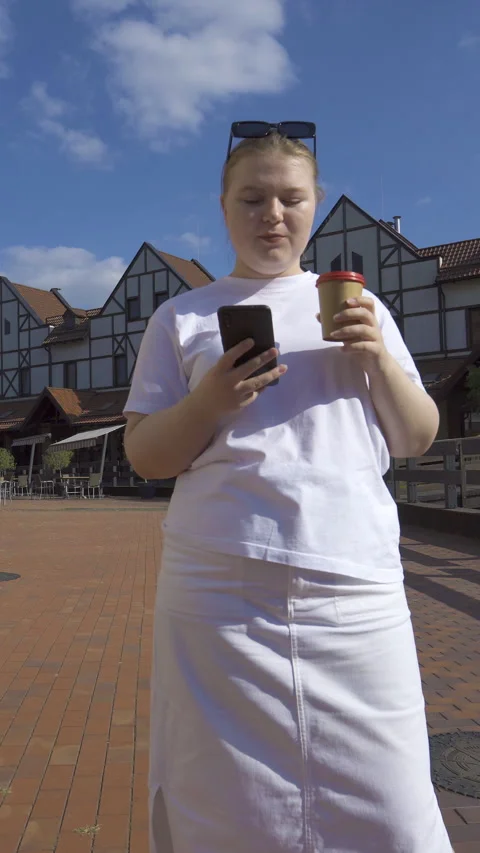 Plus size woman drinking coffee and holding phone in a sunny cottage village Stock Footage 327394980