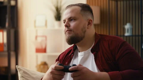 Plus-size young man enjoys playing video games, sitting on the couch alone Video stock 299621493