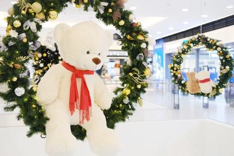 Plush teddy bear wearing red scarf hangs from festive wreath adorned with Stock Photos