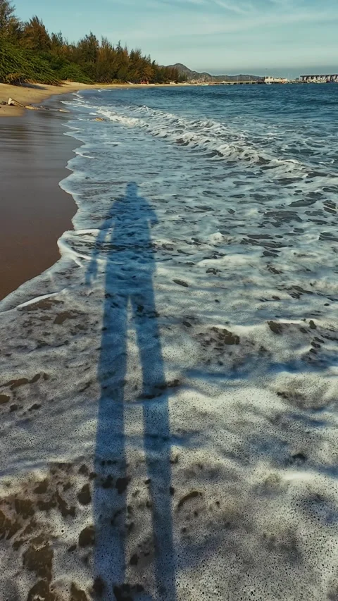 Point of View follows long shadow of person walking along sandy beach as waves Stock Footage 326364833