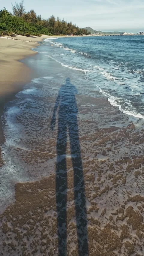 Point of View long shadow of person walking along sandy beach as waves roll in Stock Footage 326364811