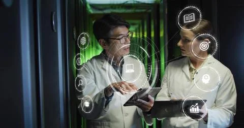 Pointing two technicians in lab coats examining tablets by server racks with Stock Photos