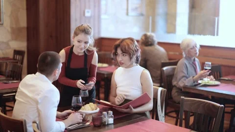 Polite female server helping young with choice in warmly decorated restaurant Stock Footage 341427776