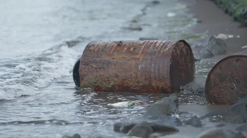 Polluted beach with rusty oil drums washing up on rocky shoreline near water Video stock 328026720
