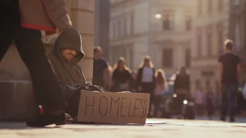 Poor homeless man sitting on street with sign people walk around sunshine Stock Footage