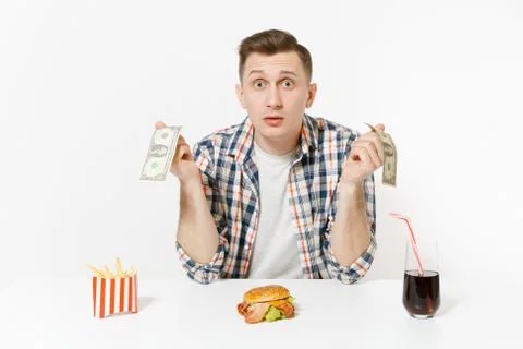 Poor sad man sitting at table with two dollar bills, burger, french fries, co Foto stock
