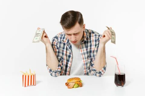 Poor sad man sitting at table with two dollar bills, burger, french fries, co Foto stock