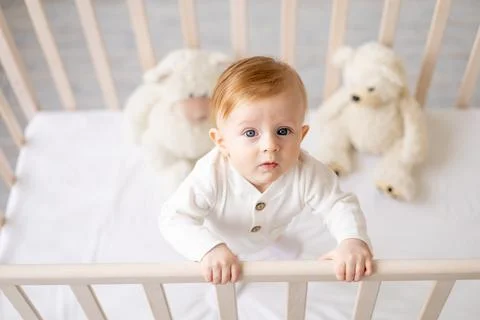 Portrait of a 6-month-old blond boy standing in a crib in a bright children's Stock Photos