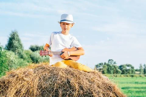 Portrait of barefoot boy in hat on haystack in field. Playing small guitar Stock Photos