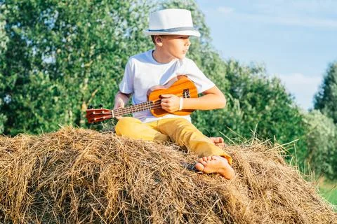 Portrait of barefoot boy in hat on haystack in field. Playing small guitar Stock Photos