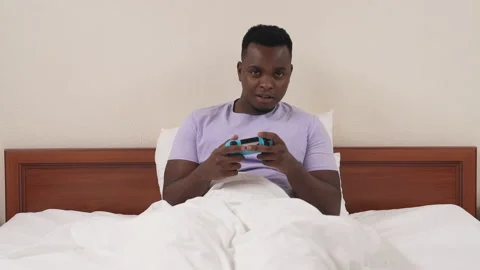 Portrait of black man lying in bed and playing console. Caucasian woman's legs Stock Footage 136586504