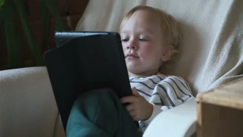 Portrait of a blond boy looking at a tablet while sitting in a chair Stock Footage 237708617