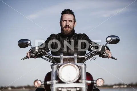 Portrait of confident biker sitting on motorcycle against sky Stock-Fotos